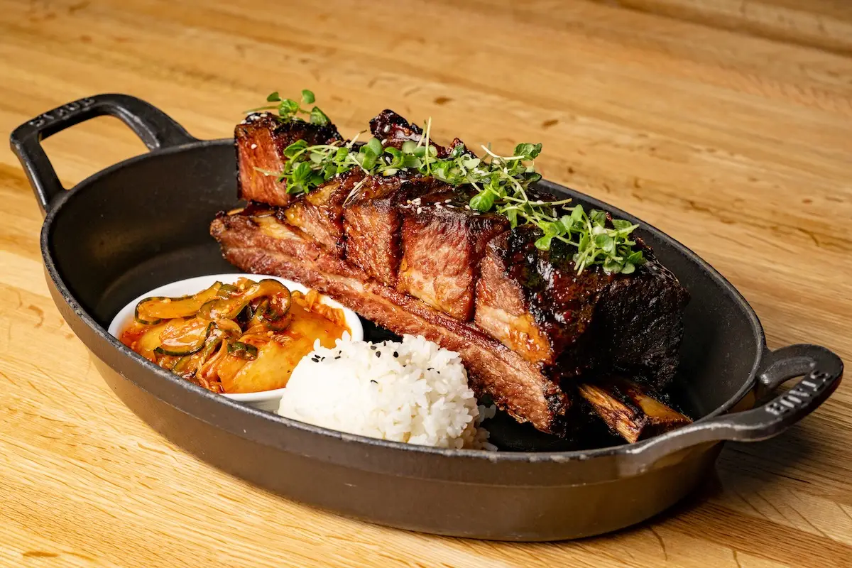 A black cast iron dish holds a large, cooked, bone-in short rib garnished with microgreens. Next to the rib are portions of white rice and what appears to be a creamy, orange-hued side dish with sautéed mushrooms—a perfect Memorial Day BBQ spread placed on a wooden surface.