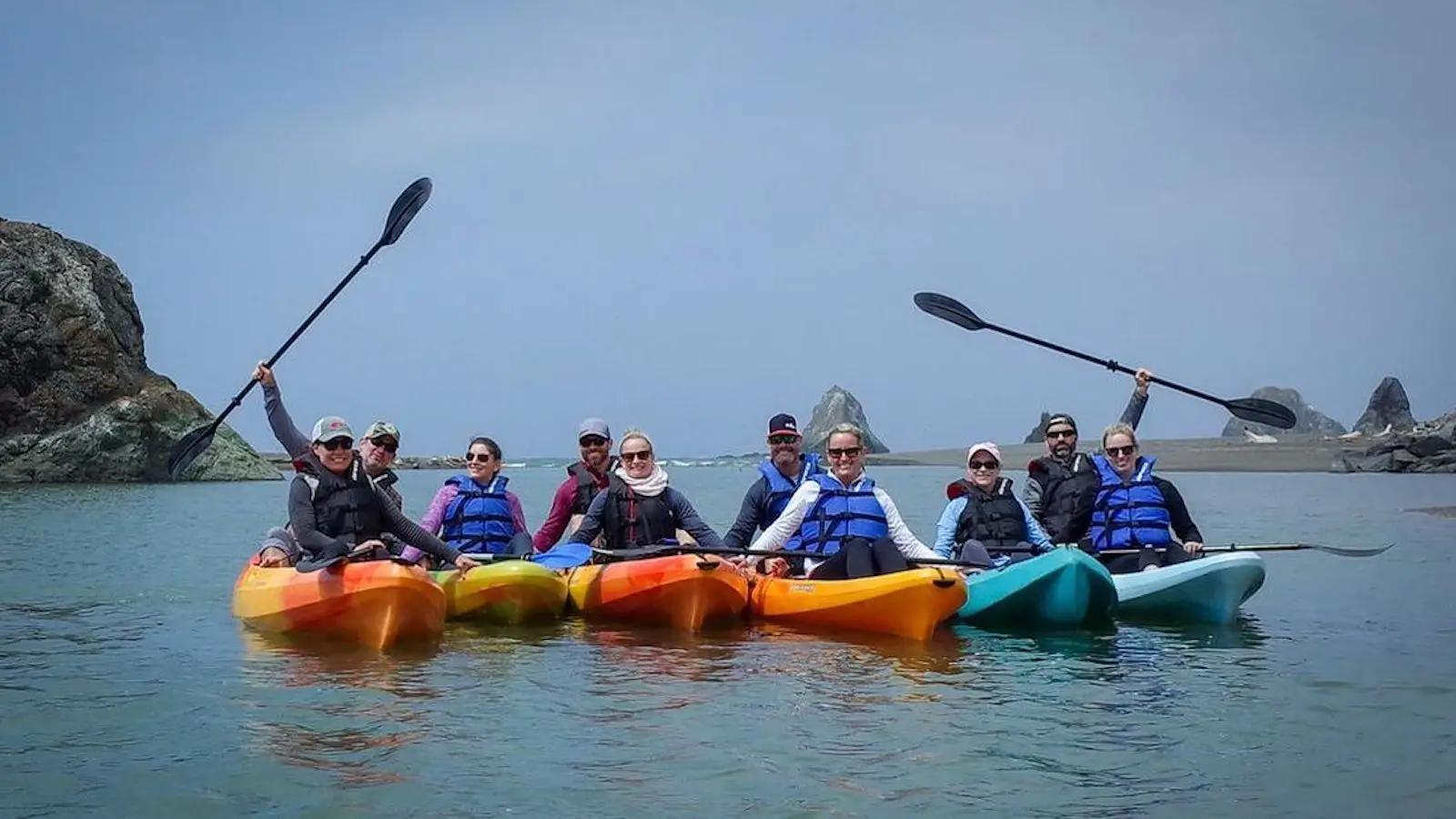 A group of people in colorful kayaks, wearing life jackets and holding paddles, are gathered on a calm body of water with rocky formations in the background. The sky is clear, and everyone appears to be enjoying one of the top activities in Sonoma.