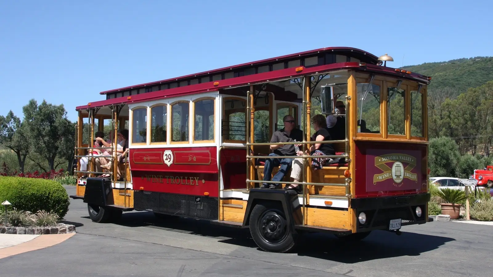A red and gold trolley with people riding on it is moving down a paved road surrounded by trees and greenery. The trolley, labeled "Wine Trolley," has an open-air design with wooden seats. It is a sunny day with a clear blue sky—a perfect setting for one of the top activities in Sonoma.