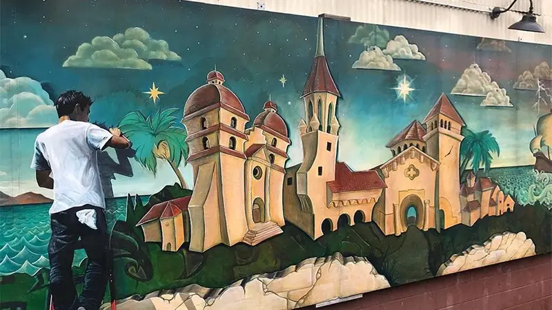 An artist paints a mural depicting a seaside village with domed buildings and church towers, reminiscent of Southern California Missions. The scene includes lush greenery, cloudy skies, and a starry night. The artist stands on a chair, adding details to a palm tree on the left side of the mural.