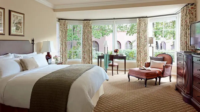 A spacious, elegant bedroom with a large bed, adorned with white linens and a green throw. The room features a sitting area with a chair and footrest, reminiscent of Southern California Missions. A desk by large bay windows offers views of greenery and a pink building outside, complemented by wooden furniture.