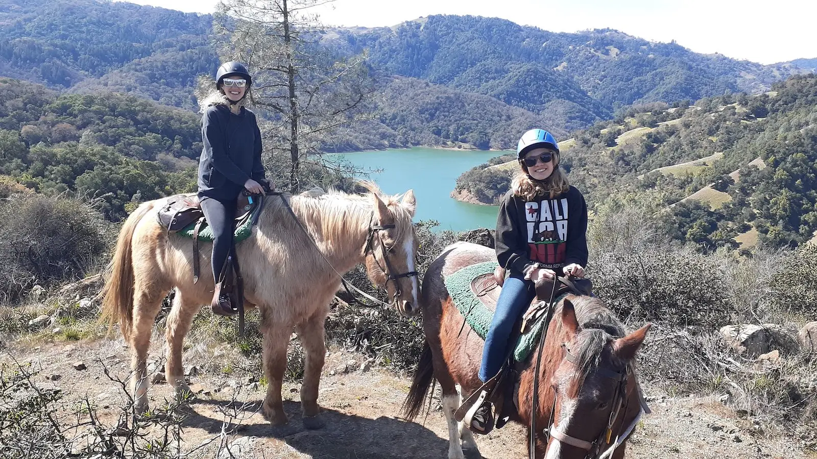 Two people are riding horses on a dirt trail with a scenic background of mountains and a lake. Both are wearing helmets and casual attire. One rider is on a light-colored horse, and the other is on a darker horse. The landscape features greenery, a clear sky, and captures one of the top activities in Sonoma.