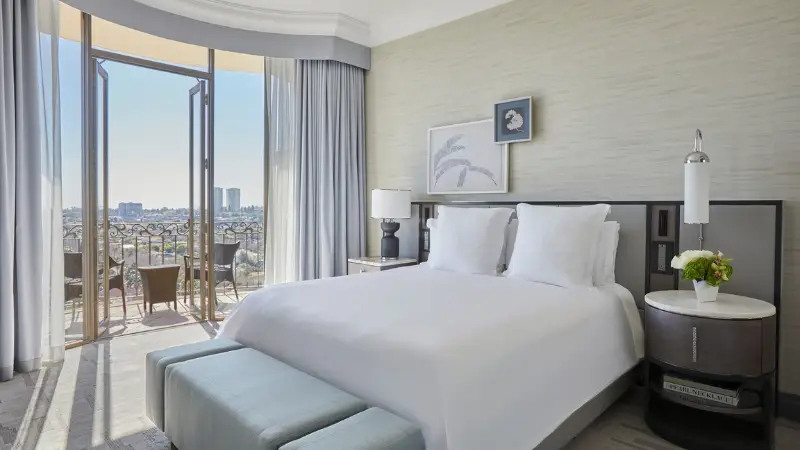 A modern hotel room features a large bed with white linens, flanked by two nightstands. A painting hangs above the bed. Glass doors open to a balcony with chairs, offering a view of a cityscape under a clear sky. Light gray curtains frame the doors—truly embodying The Ultimate Los Angeles Guide experience.