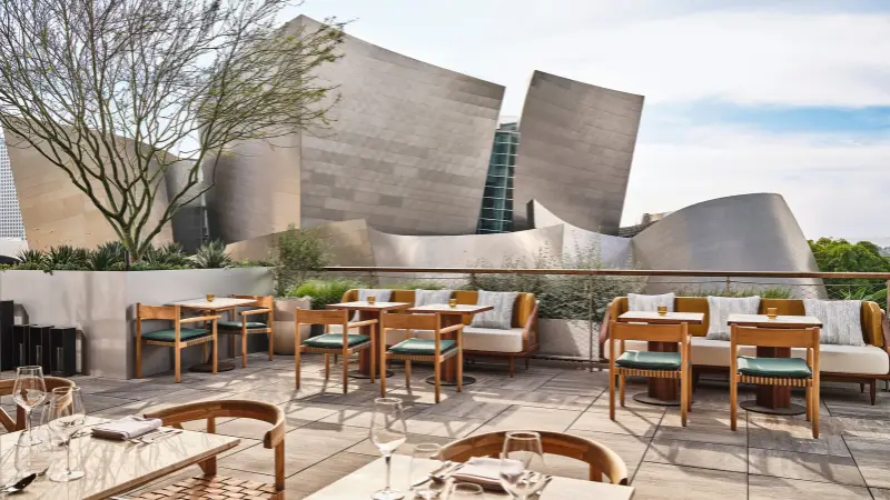 A modern outdoor patio with wooden furniture and cushioned seating is set up for dining. In the background stands a striking, contemporary building with curving metallic walls. Lush green plants dot the patio, and the sky is clear—just like you'd find in The Ultimate Los Angeles Guide.