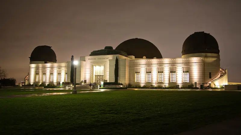 Night view of the Griffith Observatory, a grand building with two domes and a central tower, illuminated with warm lights against the dark sky. A well-maintained lawn and pathway are visible in the foreground, making it a must-see spot featured in The Ultimate Los Angeles Guide.