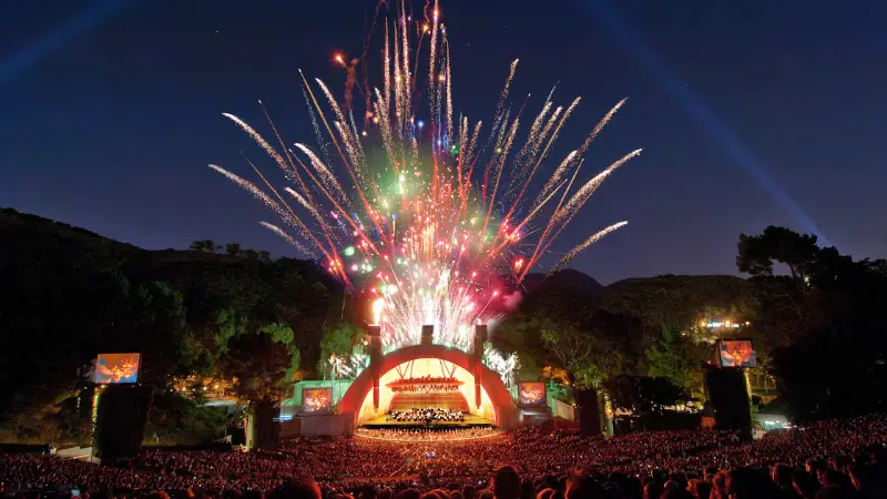 A vibrant outdoor concert at night with a semicircular stage illuminated by colorful fireworks exploding overhead. The area, highlighted in *The Ultimate Los Angeles Guide*, is surrounded by trees, and a large crowd watches the performance. Multiple screens display the stage view for the audience.