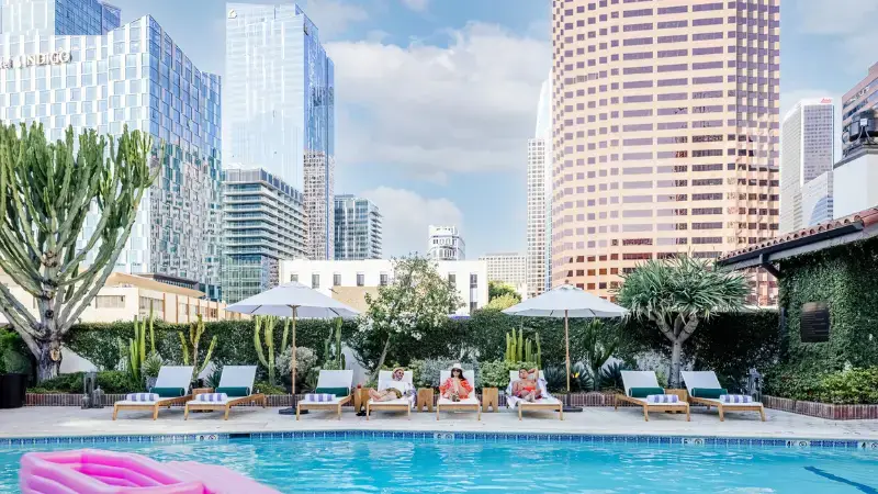 A rooftop pool area in a city with several tall buildings in the background. People are lounging on sunbeds under white umbrellas near the pool, which has a pink inflatable float. The area is surrounded by greenery, including trees and large cacti—truly a scene out of The Ultimate Los Angeles Guide.