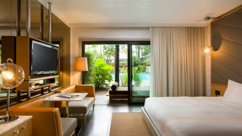 A modern hotel room with a king-sized bed, a flat-screen TV, and a cozy seating area. The room has a large glass door leading to a balcony with a view of lush greenery and a swimming pool. Warm lighting and contemporary decor create an inviting atmosphere, perfect for following *The Ultimate Los Angeles Guide*.