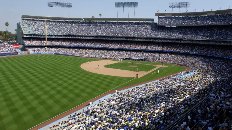 A packed baseball stadium filled with spectators enjoying a live game, as featured in The Ultimate Los Angeles Guide. The field boasts a well-maintained green outfield and a dirt infield. Players are positioned on the field, with one team at bat and the other defending, all under bright, clear skies overhead.
