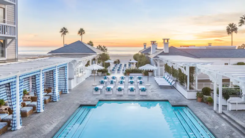 A luxurious seaside resort, highlighted in The Ultimate Los Angeles Guide, features a clear blue swimming pool surrounded by lounge chairs with blue and white striped towels. Elegant white buildings and cabanas with umbrellas are nearby, set against a scenic sunset over the ocean with palm trees in the distance.