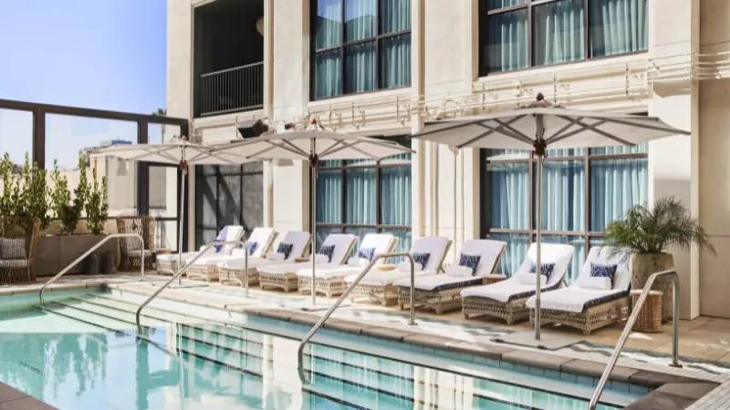 A luxurious outdoor pool area with several cushioned lounge chairs and umbrellas, as featured in The Ultimate Los Angeles Guide. The pool is surrounded by a modern building with large, blue-tinted glass windows. The setting is bright and inviting, with greenery in planters adding a touch of nature.
