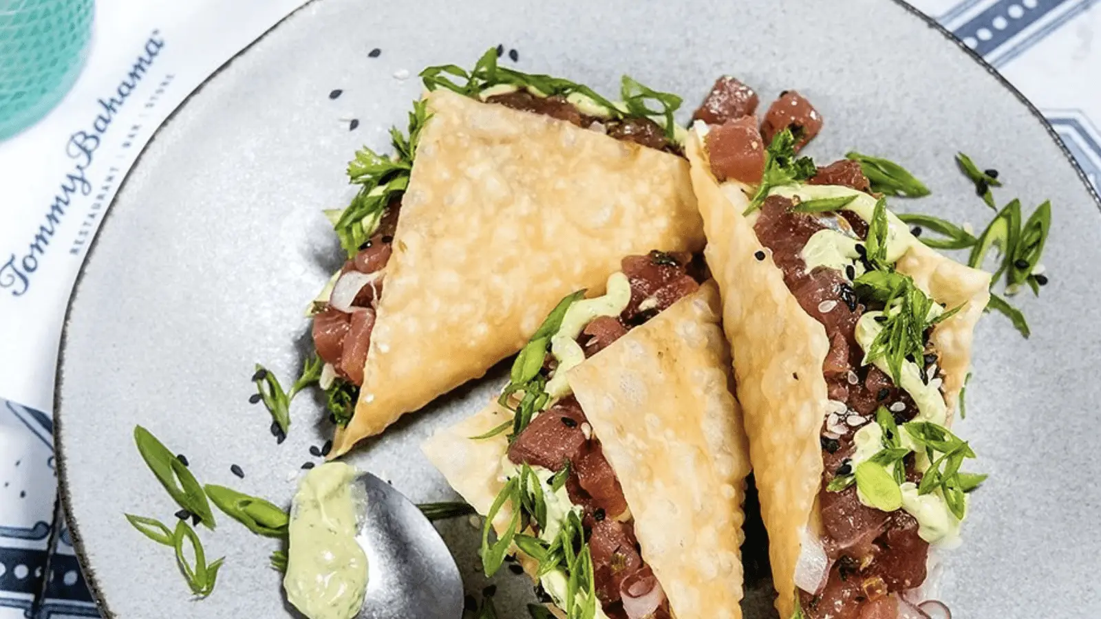 A plate with three triangular wonton tacos filled with diced raw tuna, green onions, and sesame seeds, garnished with greens. A dollop of wasabi mayo sits beside them at the best dinner spot in Kihei Wailea. Part of a Tommy Bahama restaurant napkin and a drink are partially visible in the background.