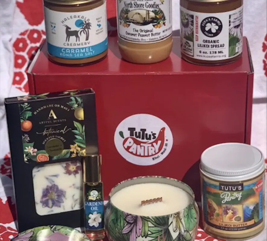 A delightful assortment of gourmet and artisanal products on a red box featuring the "Tutu's Pantry" logo. Perfect as Valentine's Day gifts from Hawai'i, it includes caramel sauce, coconut peanut butter, lilikoi spread, a floral-scented candle, gardenia oil, and a box of chocolates with a botanical design.
