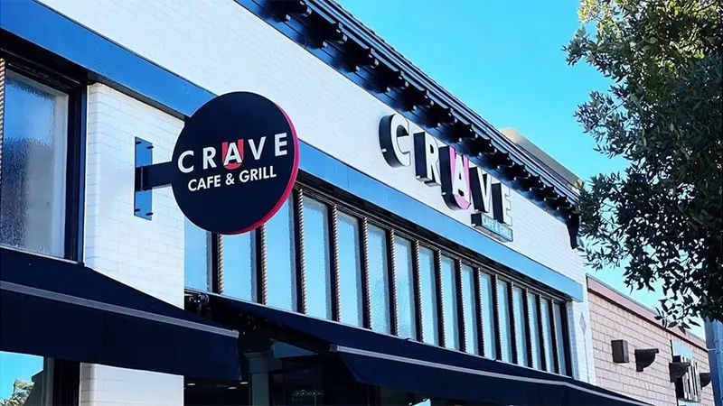 A street view of the exterior of Crave Cafe & Grill with a black circular sign hanging above the entrance on the left and a large white sign with bold letters on the building facade. The sky is clear blue, and some tree branches are on the right, reminiscent of Southern California Missions' serene landscapes.