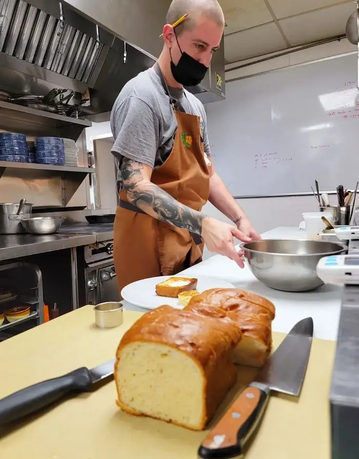 A person with a shaved head and a tattooed arm, wearing an apron and face mask, is preparing food in a commercial kitchen. In the foreground, a loaf of bread is partially sliced on a cutting board beside a knife, seemingly ready for the upcoming Memorial Day BBQ.