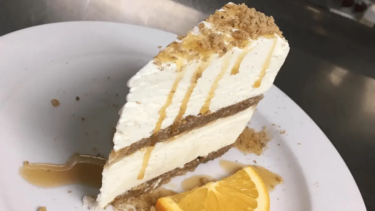 A slice of layered cheesecake with a crumb crust is served on a white plate in the heart of Wailea. The cheesecake, celebrated as one of the best desserts in Maui, is drizzled with a light syrup and garnished with a slice of orange. Crumbs are delicately sprinkled around the plate.