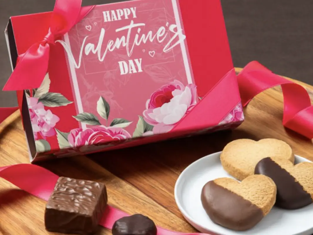 A red and pink Valentine's Day gift box with a floral design and ribbon sits on a wooden surface. In front of the box, there are chocolate-covered heart-shaped cookies on a white plate, along with square and round chocolates, perfect for those seeking unique Valentines Day Gifts from Hawai'i.
