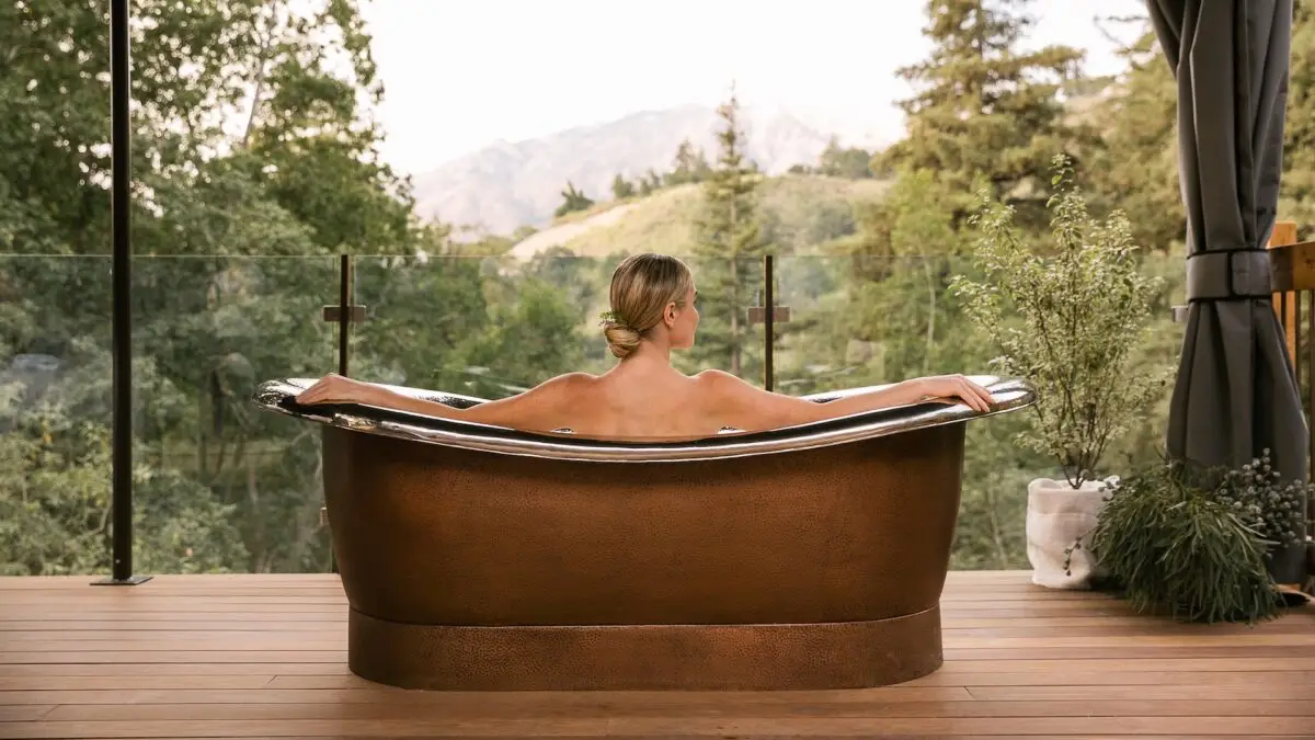 A person with blonde hair is relaxing in a large, elegant bronze bathtub on a wooden deck. Surrounding the deck are lush green trees and plants. In the background, there are scenic mountains and a clear sky, creating a tranquil outdoor setting reminiscent of Big Sur spa and wellness hotels.