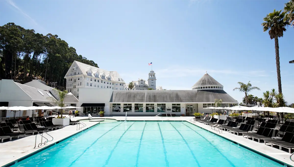 A large rectangular swimming pool with lounge chairs and umbrellas on both sides. In the background, there are white buildings with triangular roofs, including one with a dome-like structure. Trees and a clear blue sky complete the serene setting, typical of California eco hotels.