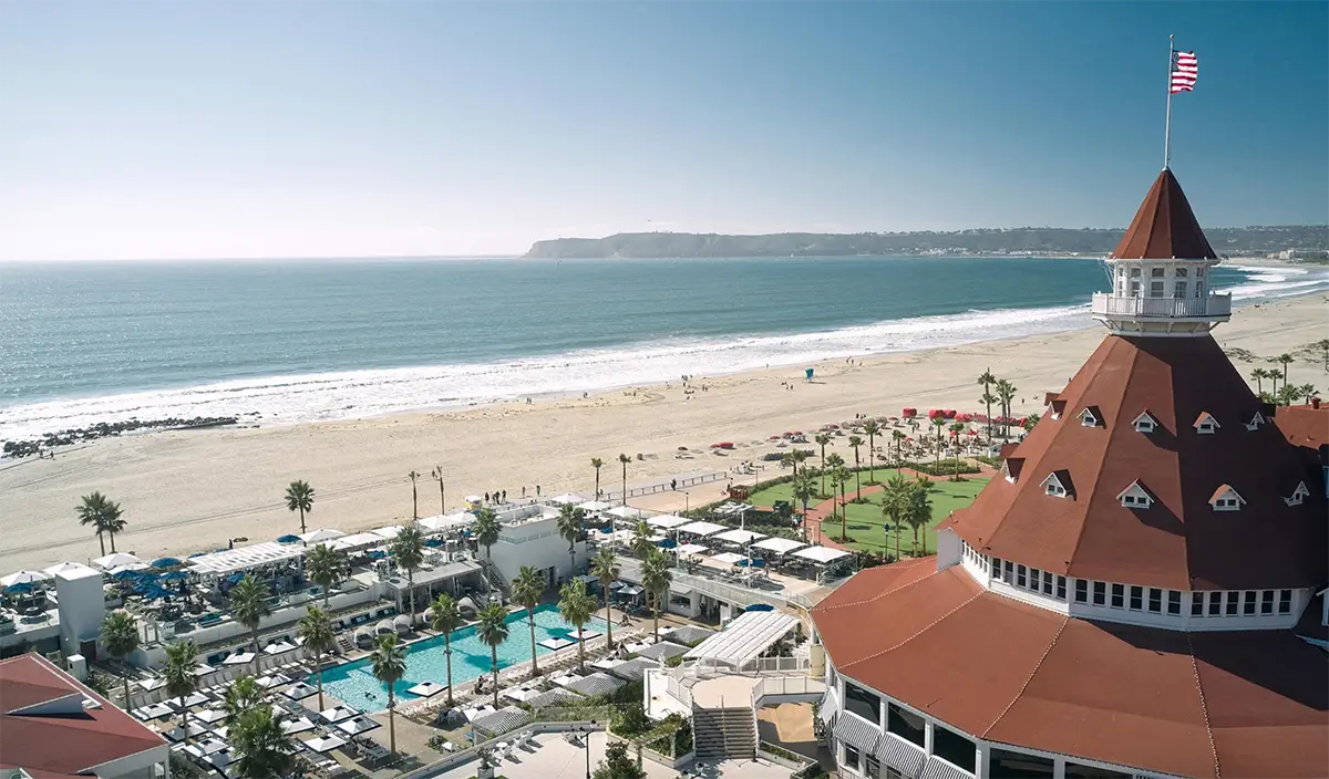 An aerial view of one of the best group hotels in San Diego, featuring a large, red-roofed building with a pointed tower. The property includes a pool area, palm trees, and beachgoers enjoying a sandy beach beside a calm ocean under a clear blue sky. An American flag flies atop the tower.