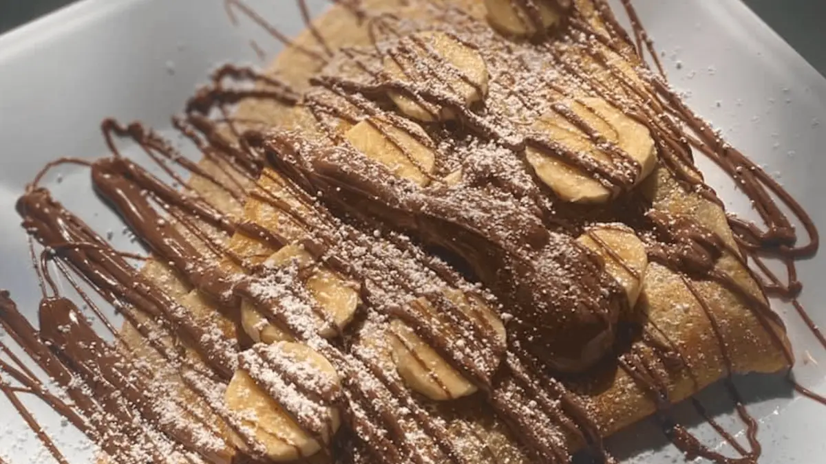 A close-up of one of the best desserts in Wailea: sliced bananas atop a folded crepe, generously drizzled with chocolate sauce and lightly dusted with powdered sugar. This delectable dish is served on a pristine white plate.