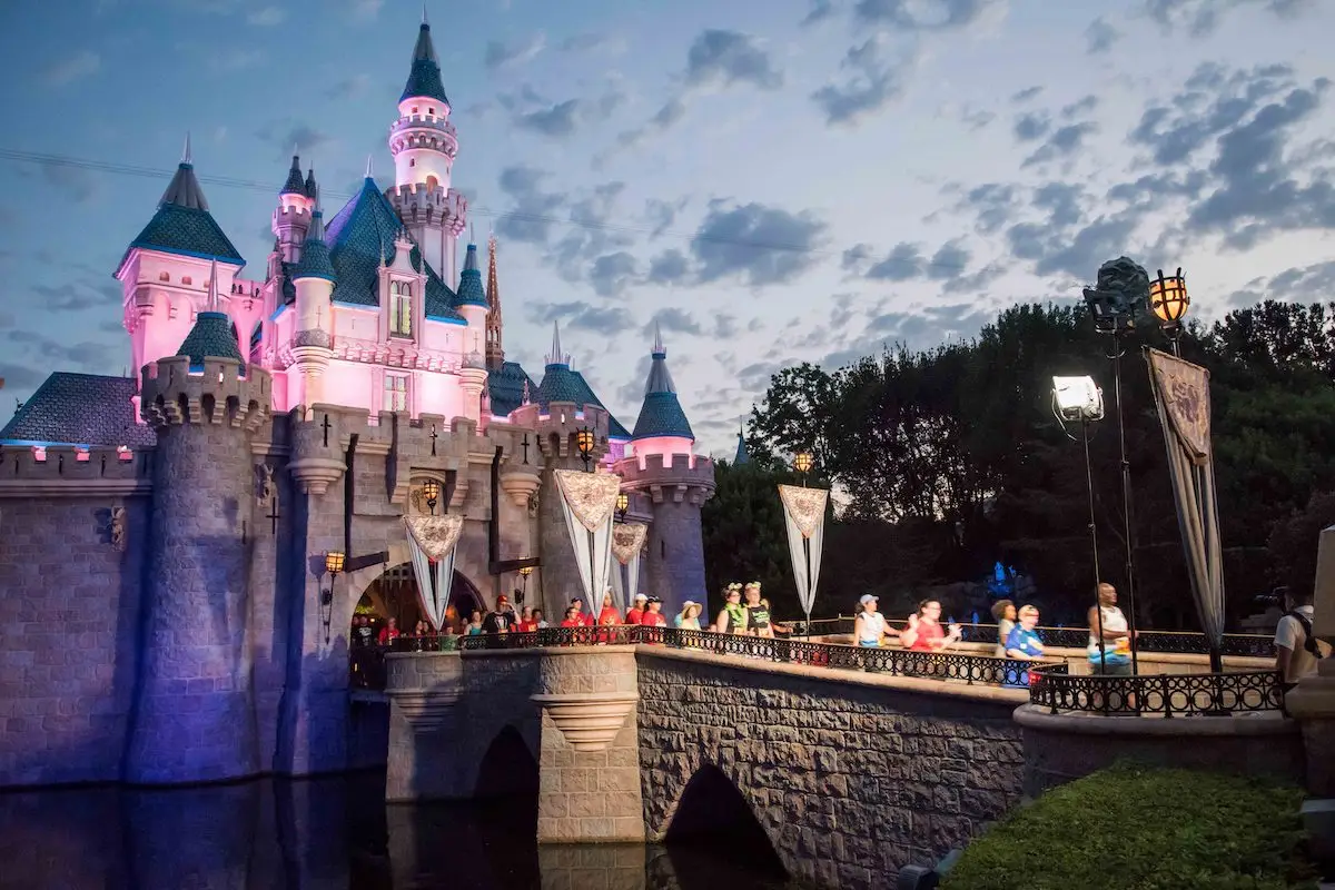 A picturesque castle with pink and blue spires is illuminated in the evening. A stone bridge crosses a body of water leading to the castle's entrance. People are walking on the bridge, and the sky is scattered with clouds as dusk approaches, setting a perfect scene for Disneyland 2024 events.