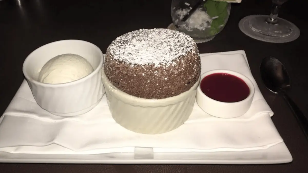 A round chocolate souffle dusted with powdered sugar, served in a white ramekin, sits elegantly on a white rectangular plate. Accompanied by a scoop of vanilla ice cream and a small bowl of red sauce, this delightful treat is one of the best desserts in Maui Wailea. A spoon rests beside the plate.