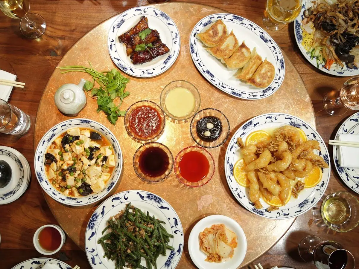A circular table set with various Chinese dishes, including dumplings, stir-fried vegetables, shrimp, tofu, and dipping sauces. Plates and glasses surround the central spread, emphasizing a communal meal arrangement reminiscent of the exquisite dining experiences at Berkeley Michelin restaurants.