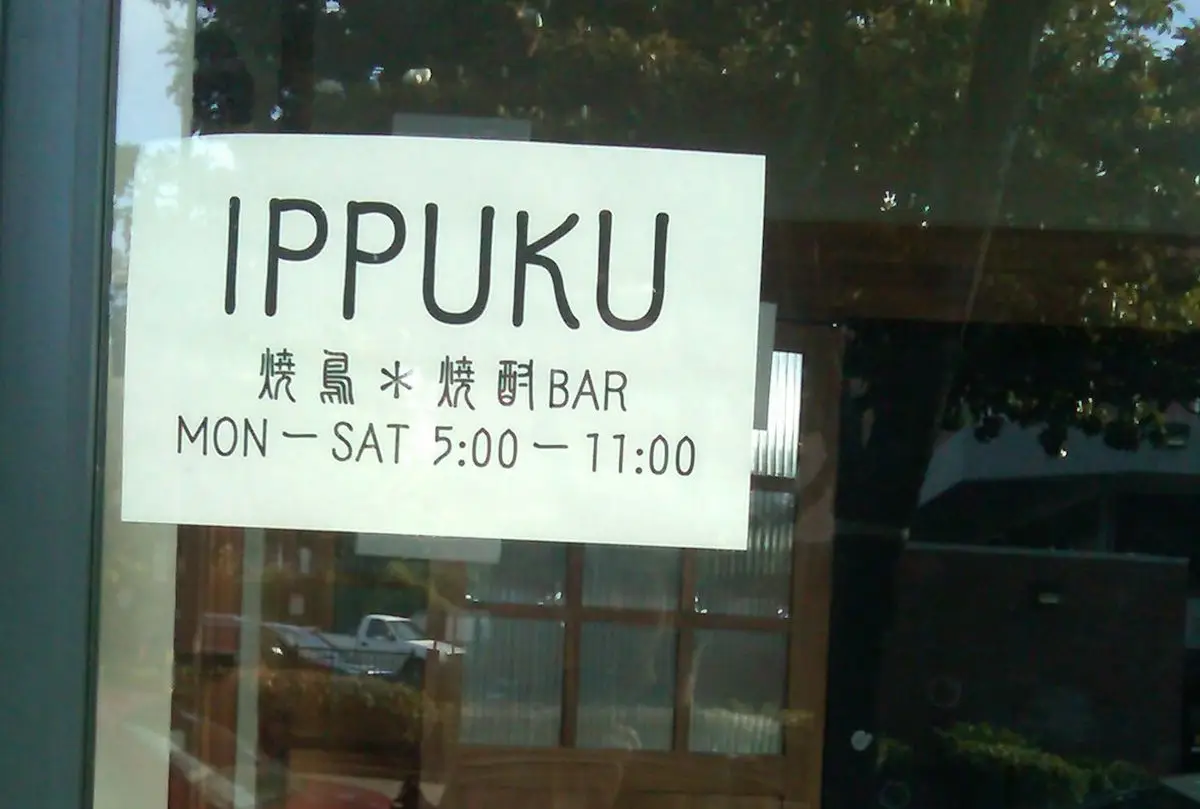 A white sign posted on a glass door reads: "IPPUKU, 焼鳥*焼酎BAR, MON - SAT 5:00 - 11:00." The background shows reflections of trees and a building, inviting patrons to experience one of Berkeley's Michelin restaurants.