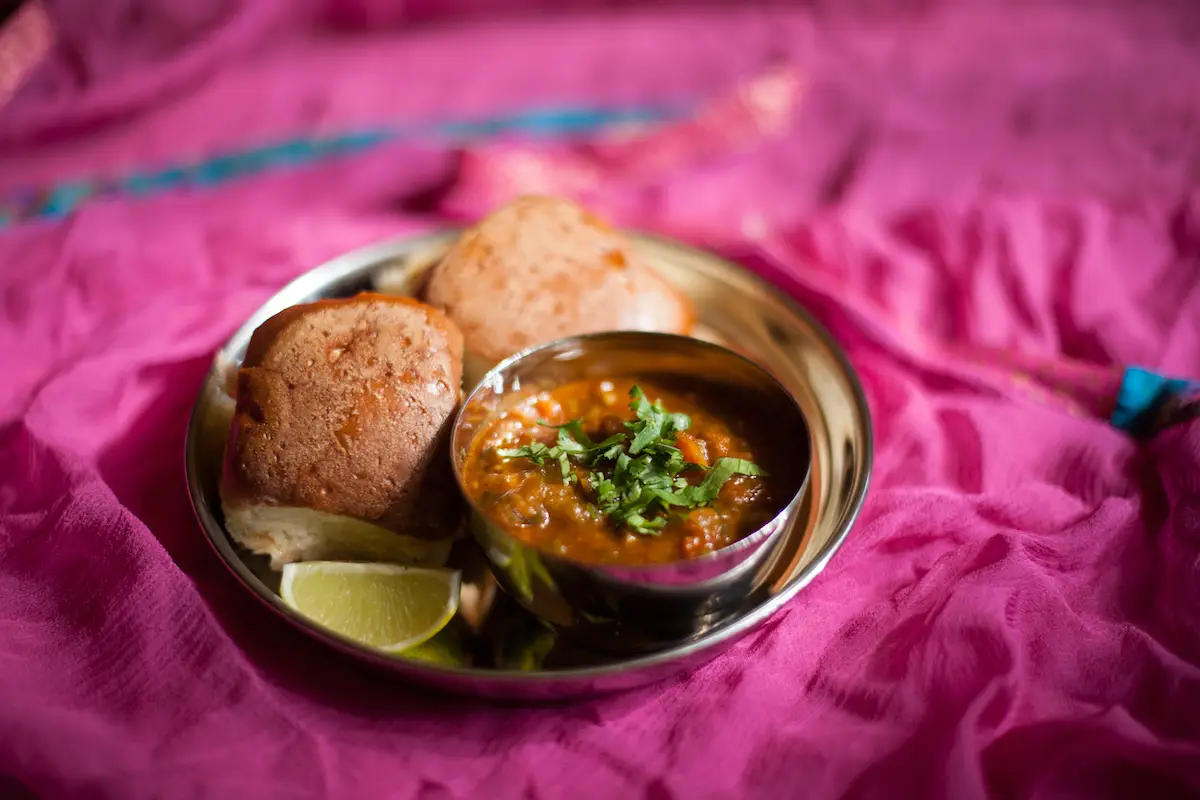 A stainless steel plate on a pink cloth holds three pieces of bread, a small bowl of vegetable curry, and a lime wedge. The curry is garnished with fresh coriander leaves, reminiscent of the exquisite presentations found in Berkeley Michelin restaurants.