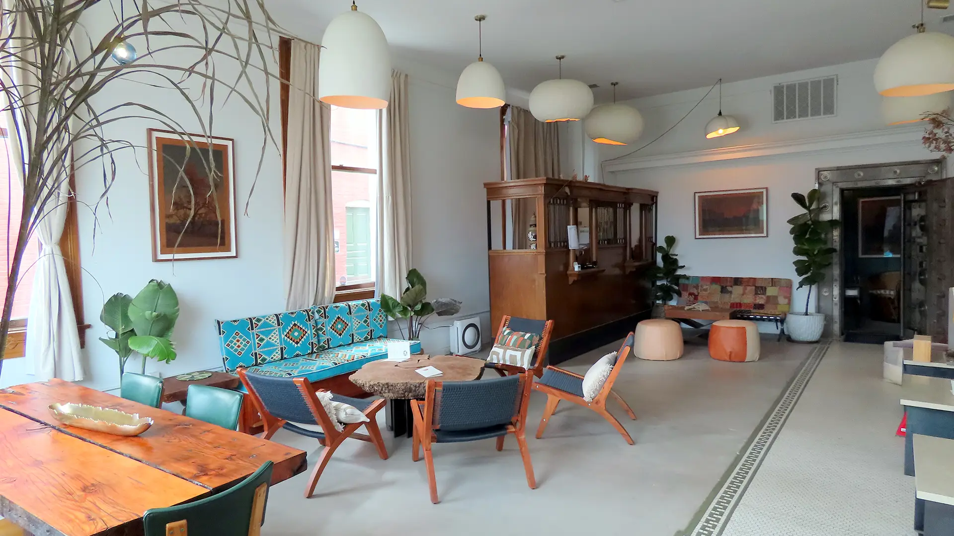 A modern living room with a mix of contemporary and vintage furniture, reminiscent of Sonoma tasting rooms. Pendant lights hang from the ceiling, illuminating a patterned blue sofa with wooden chairs, a wooden dining table with chairs, and various plants and framed artwork adorning the walls.