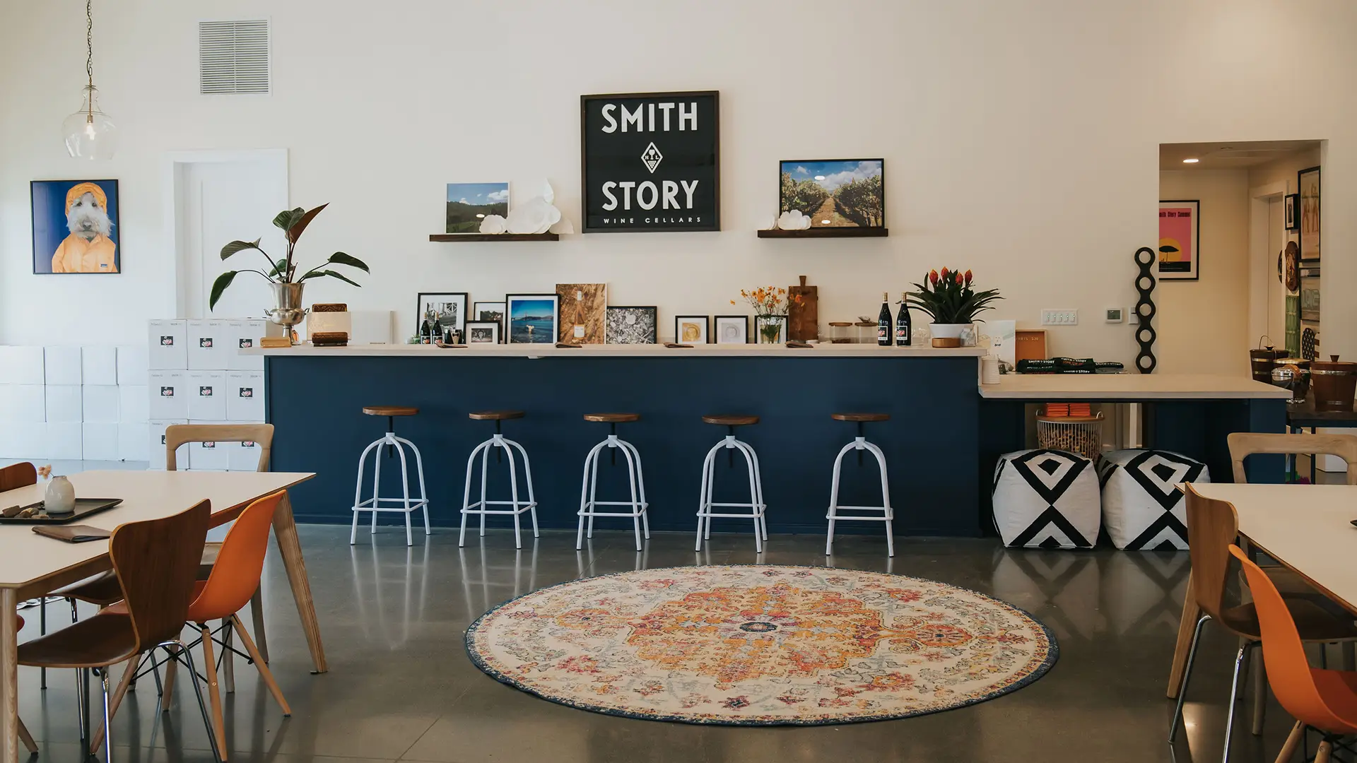 A modern cafe interior with a blue bar counter lined with five stools, reminiscent of the chic Sonoma Tasting Rooms. The back wall features shelves displaying various framed pictures and decor items, with a "Smith Story Wine Cellars" sign in the center. A round, colorful rug and wooden chairs complete the cozy setting.