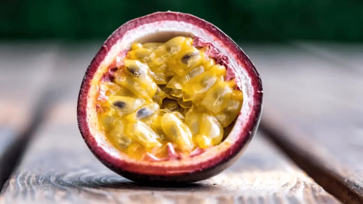 A close-up of a halved passion fruit displayed on a wooden surface, reminiscent of the best desserts in Maui Wailea. The fruit's outer skin is purple, and the inside is filled with juicy, yellow seeds surrounded by a gelatinous pulp. The background is blurred, focusing attention on the fruit.