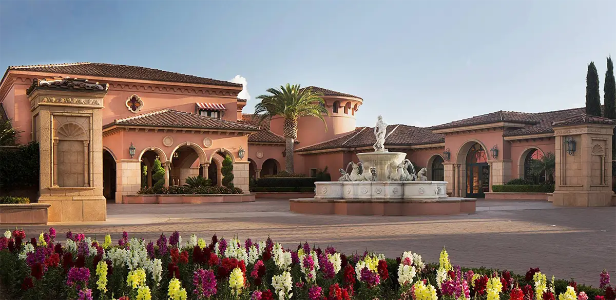A luxurious, eco-friendly Mediterranean-style building with terracotta rooftops and pink walls, featuring multiple archways and a large central fountain adorned with statues. Colorful flowers line the foreground, and a tall palm tree stands near the building, embodying the charm of California Eco hotels.