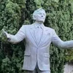 A bronze statue of a smiling man in a suit and tie with arms outstretched, resembling Tony Bennett, stands proudly. Set against a backdrop of dense, green foliage reminiscent of San Francisco's parks, the man's expression is welcoming and joyful.