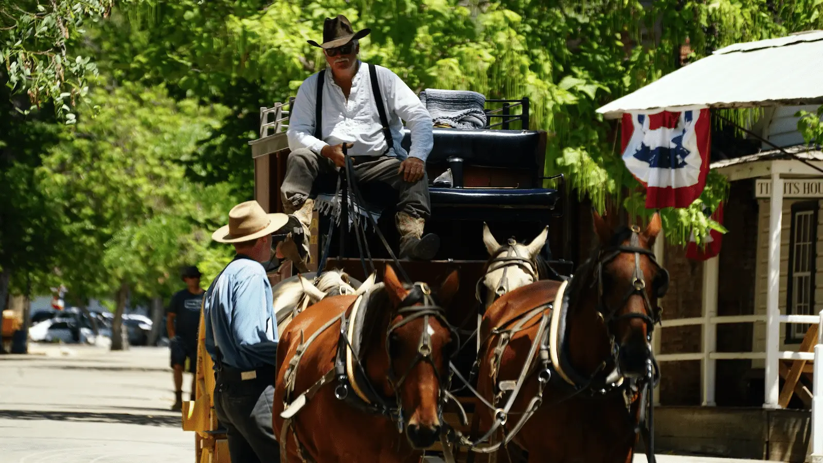 A man dressed in historical attire drives a stagecoach pulled by four horses down a tree-lined street. Another man, also in period costume, stands nearby talking to him. The scene appears to be set in California Gold Country, evoking the charm of old mining towns, with a flag hanging from a building.