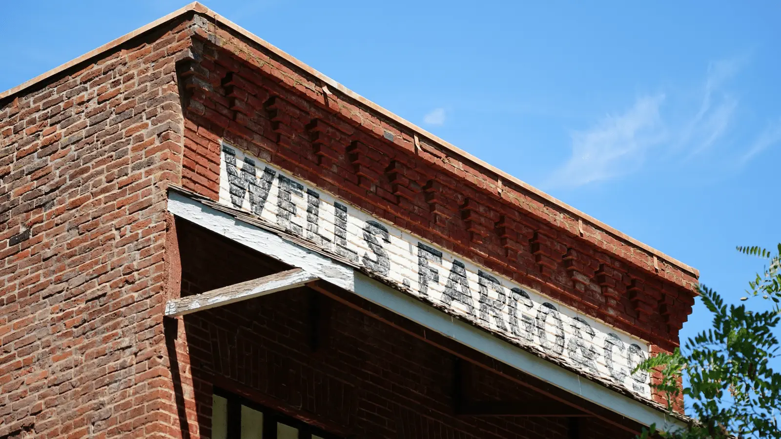 A close-up of the top corner of a red brick building with a weathered sign that reads "Wells Fargo" against a clear blue sky, reminiscent of structures in California Gold Country mining towns. The facade features distinctive brickwork patterns and a canopy with peeling paint. Green foliage is partially visible on the right.