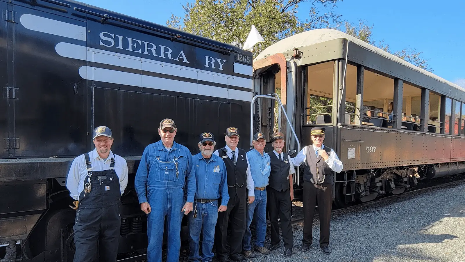 A group of seven people, dressed in train conductor uniforms and overalls, stands in front of a vintage train labeled "Sierra Ry." The black engine and connected passenger car evoke the charm of trains and museums in Northern California. Trees are visible in the background under a clear sky.