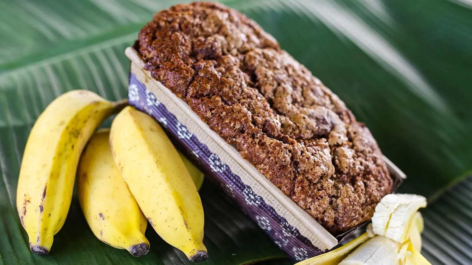 A freshly baked loaf of banana bread rests in a paper tray on a large green banana leaf. Three ripe bananas and one sliced banana lie next to the loaf, showcasing the ingredients used in the bread. The overall setup gives a tropical and homemade feel to the image, reminiscent of what to do in Hana.