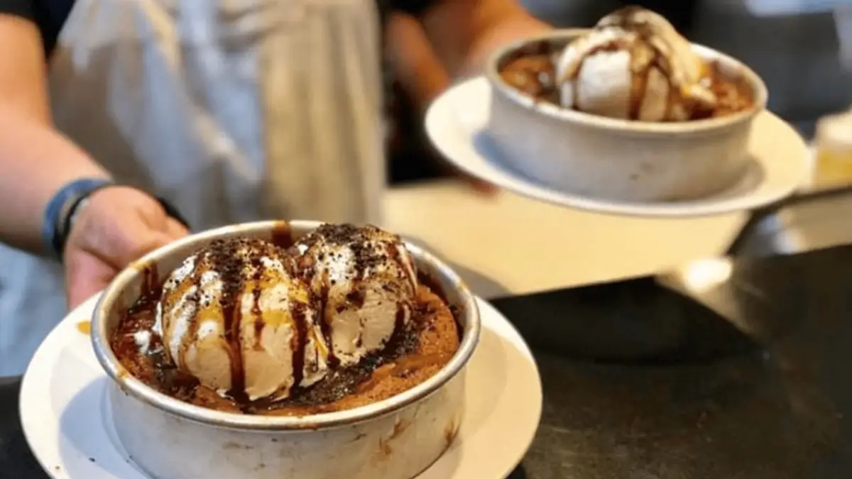 Two hands holding plates of the best desserts Maui Wailea has to offer, featuring scoops of vanilla ice cream drizzled with chocolate syrup atop a warm cookie in a metal dish. The background is slightly out of focus, highlighting the delectable treat in the foreground.