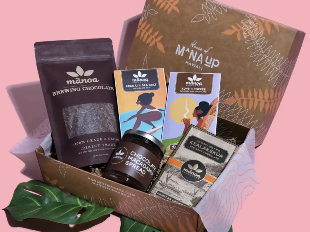 A brown box labeled "Mana Up Hawaii" contains various Hawaiian-themed products, perfect as Valentines Day Gifts from Hawai'i. Items include a package of Manoa Brewing Chocolate, a jar of chocolate macadamia nut spread, two bars of chocolate, and a coffee packet. The box sits against a pink background with leaves in front.