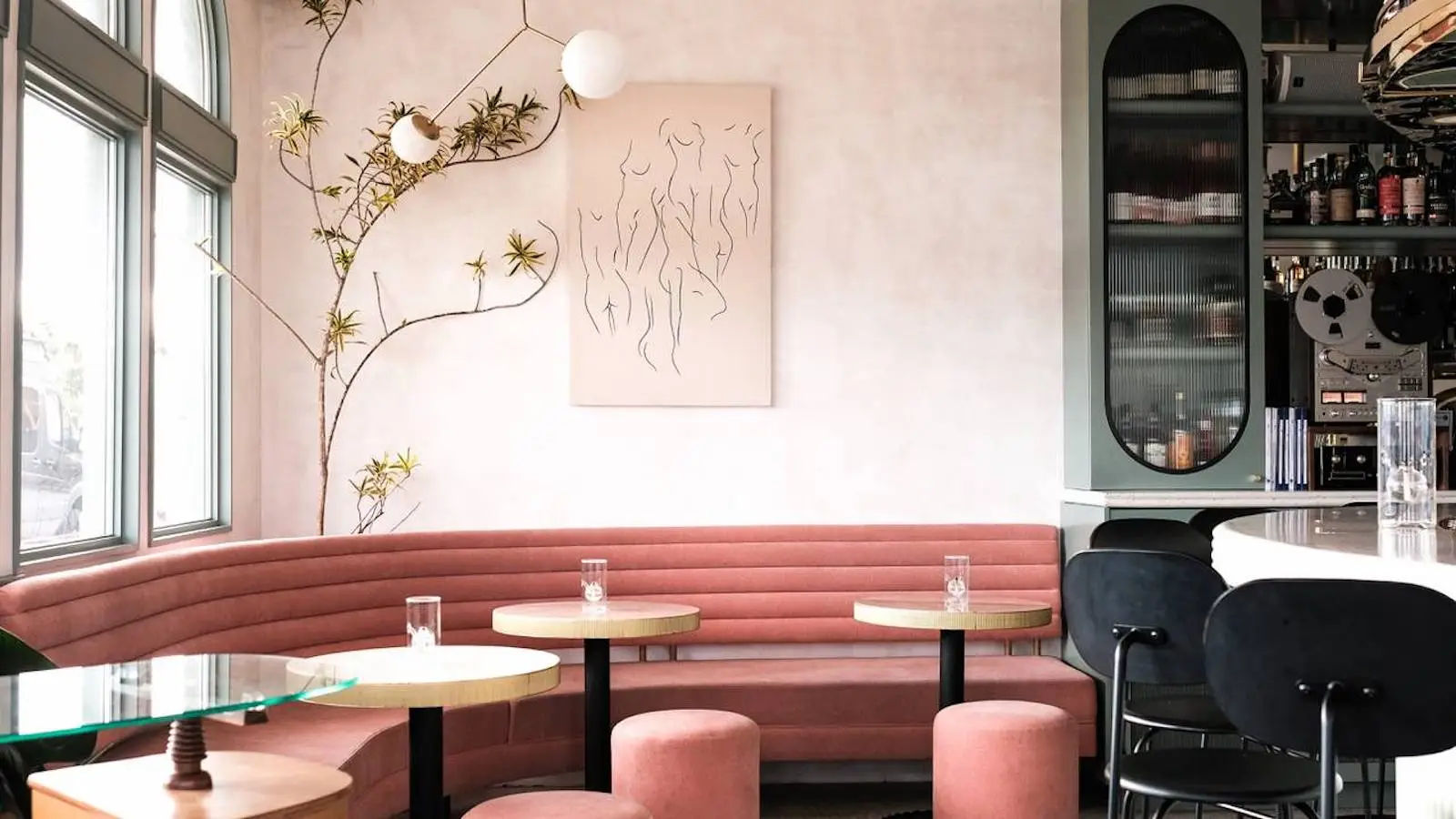 A stylish cafe interior with pink cushioned seating, round wooden tables, and pink ottomans. Walls are adorned with minimalist art and modern light fixtures. A bar area with dark stools is seen on the right, and large windows let in natural light. It's one of the best restaurants San Diego has to offer.