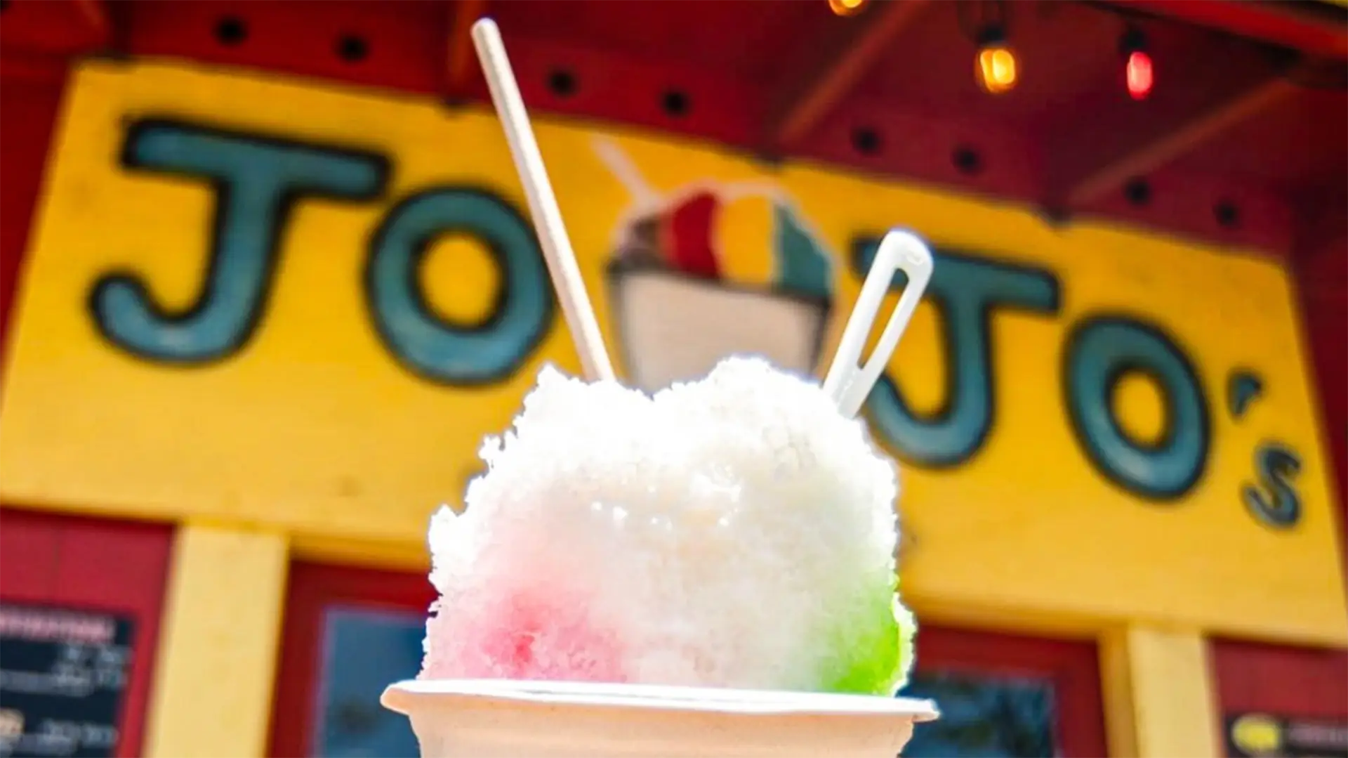 Close-up of a shave ice dessert on a white dish, featuring pink, white, and green colors, with two white spoons inserted into it. The background showcases the brightly colored yellow sign of "JoJo's," renowned for offering the best shave ice in Princeville and Hanalei.