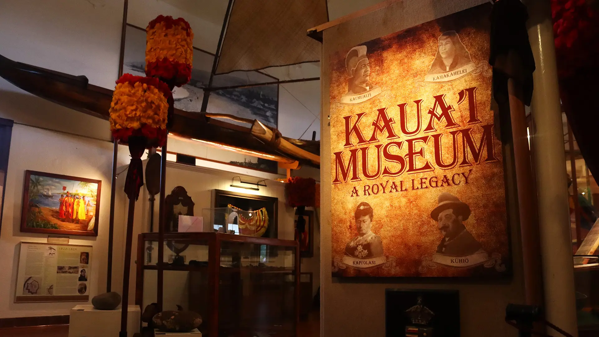A warmly lit exhibit at the Kaua'i Museum, one of the best places to visit on Kauai, features a prominent sign that reads, "Kaua'i Museum: A Royal Legacy," surrounded by historic portraits and cultural artifacts. Adjacent display cases hold various traditional items, framed pictures, and information panels.
