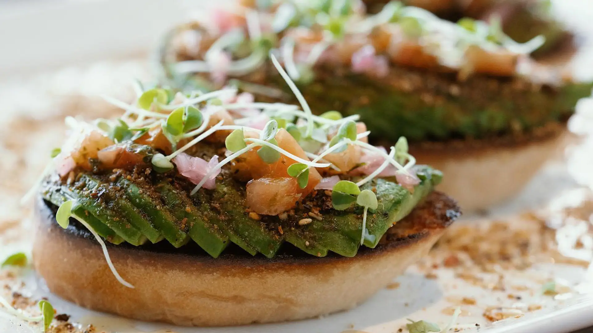 Close-up of an avocado toast on a toasted slice of bread, garnished with diced tomatoes, microgreens, and various seasonings. The toast is arranged on a white plate with bits of seasoning scattered around. Another piece of toast blurs in the background, reminiscent of the best Hawaiian food Kauai offers.