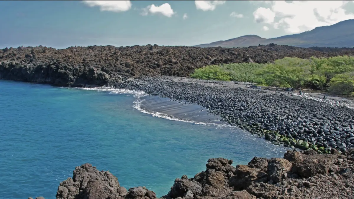 A serene rocky beach with clear blue waters and black volcanic rock formations under a partly cloudy sky, one of Maui's lesser-known beaches. Green shrubs are visible in the background along with distant mountains. Small waves gently crash onto the shore, creating a peaceful ambiance.