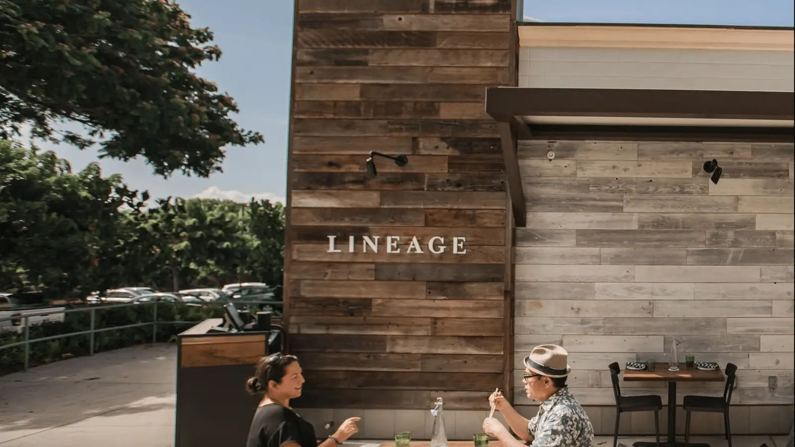 Two people are seated at an outdoor restaurant table beneath a wooden wall with the word "LINEAGE" on it. They appear to be enjoying the best dinner Kihei Wailea has to offer on a sunny day, with some greenery and parked cars visible in the background.