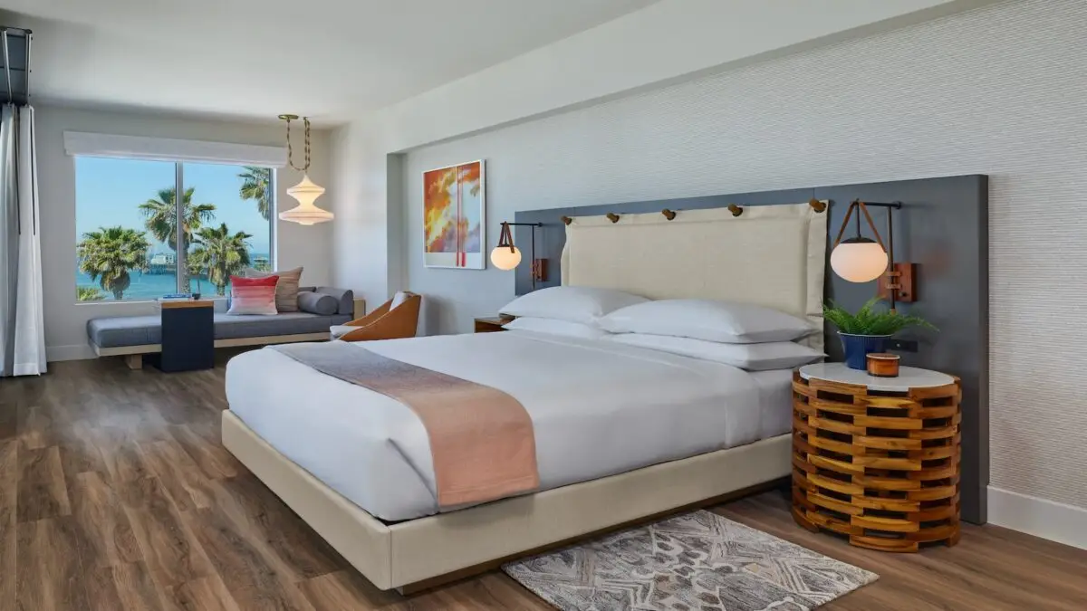 A modern hotel room in one of the best hotels San Diego has to offer, featuring a large bed with white linens and a beige headboard. There's a patterned rug on the wooden floor, a round wooden nightstand, and a lounge area with a sofa by a window showing a view of palm trees and a body of water.