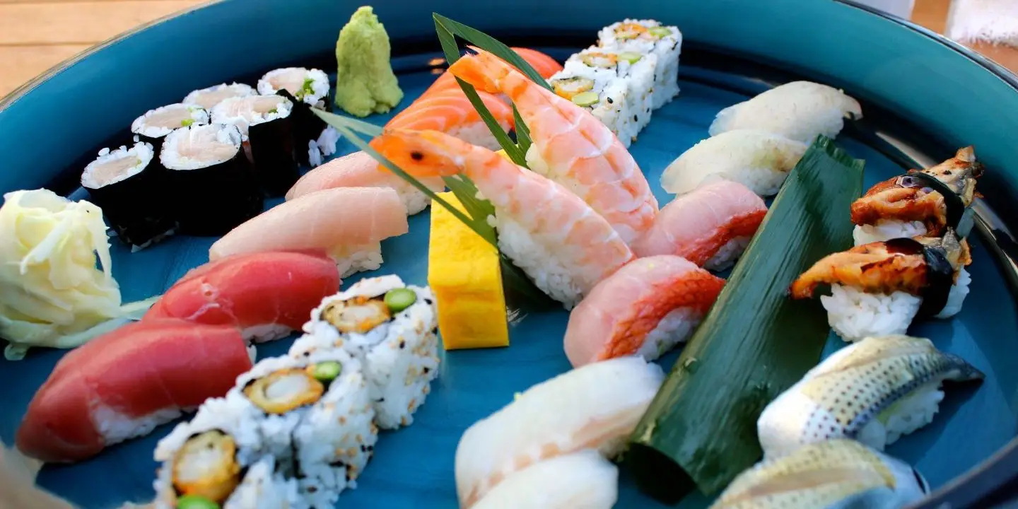 A blue platter filled with an assortment of sushi pieces, including nigiri, maki rolls, and sashimi. Various types of fish, shrimp, and vegetables are featured, along with garnishes of pickled ginger and wasabi—the best dinner you'll find in Kihei or Wailea.