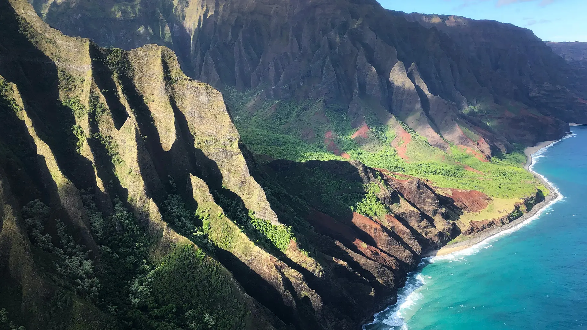 A stunning aerial view of the Na Pali Coast on Kauai, Hawaii, one of the best places to visit on Kauai. Towering cliffs with lush greenery and dramatic shadows cascade down to meet the turquoise waters of the Pacific Ocean under a bright blue sky.